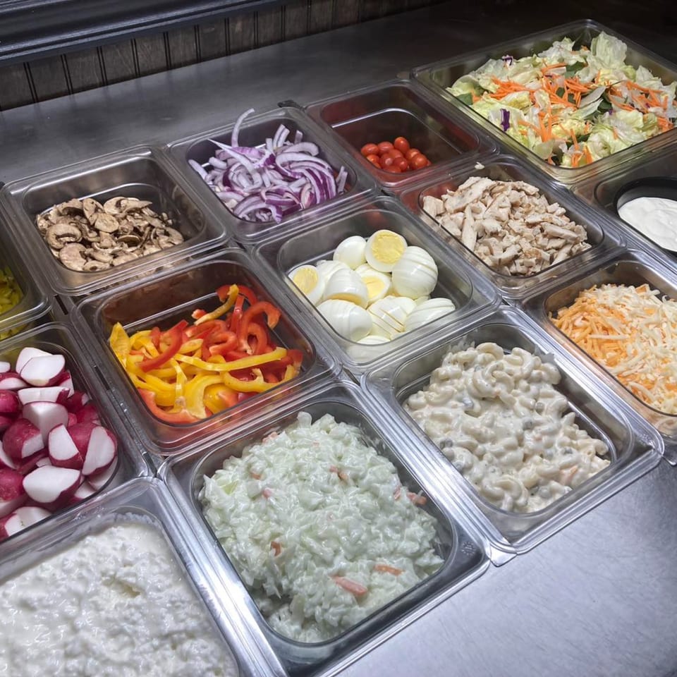 A salad bar with various toppings including vegetables, meats, and dressings in metal containers.