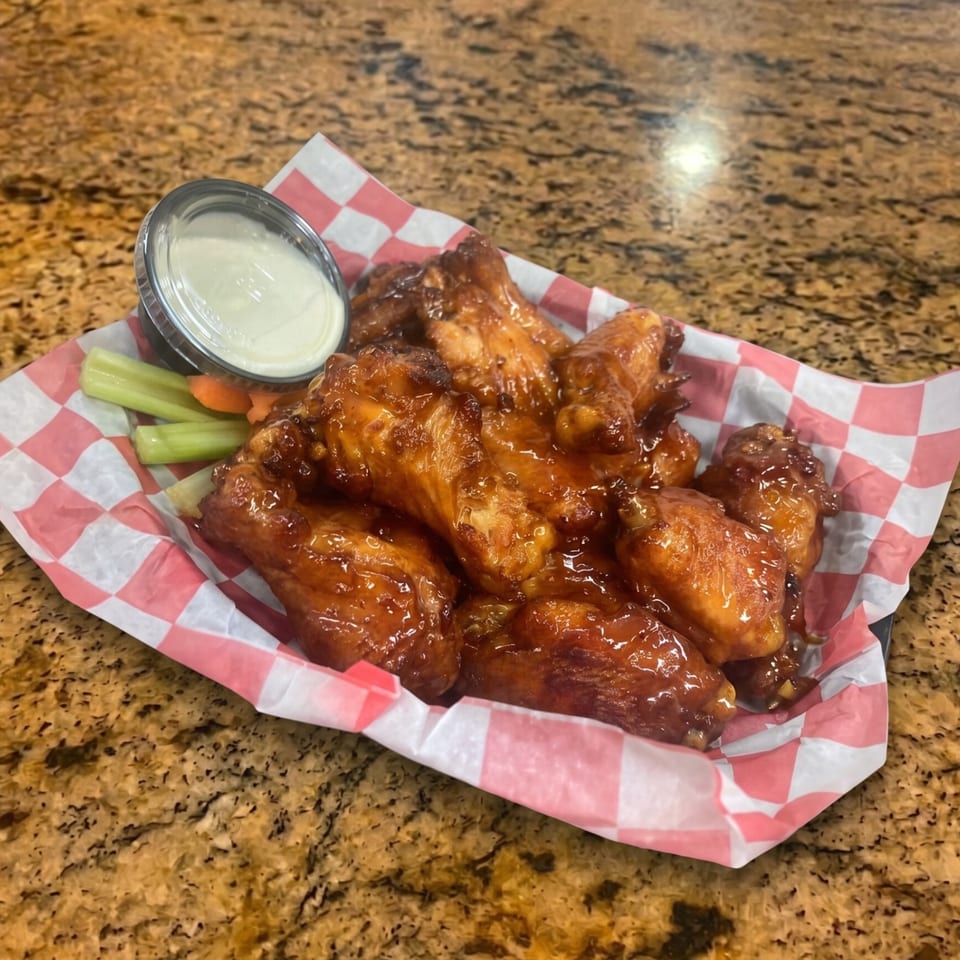 A basket of glossy, sauced chicken wings with celery, carrots, and ranch dressing on a checkered liner.