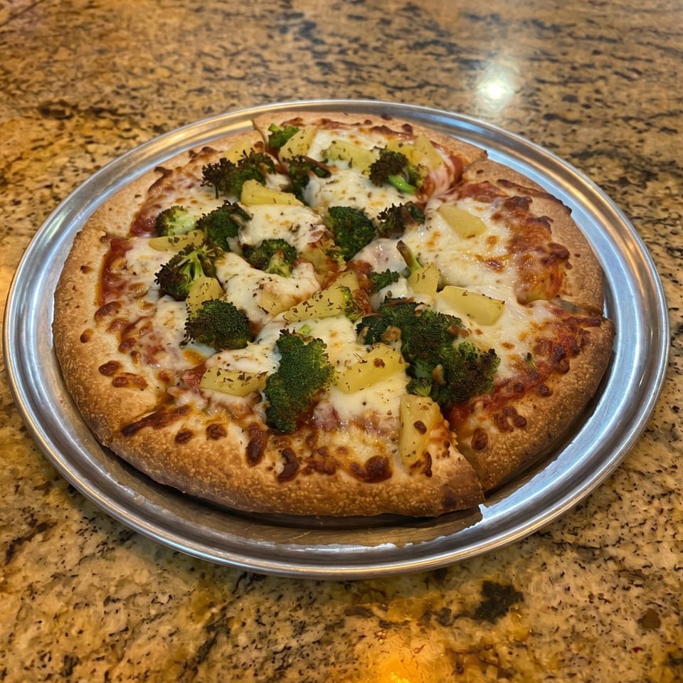 A whole pizza with broccoli and pineapple toppings on a metal tray, on a granite countertop.